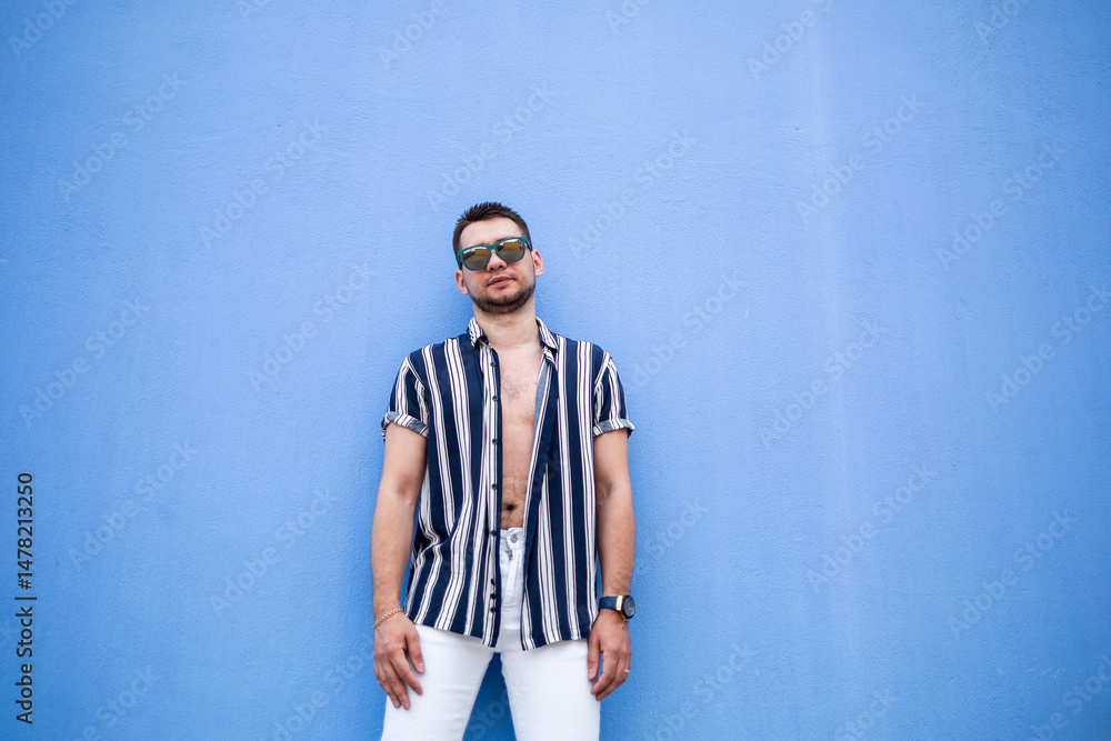 A stylish young man poses confidently in a striped shirt against a blue wall, showcasing modern outdoor lifestyle with chic accessories and sunglasses, embodying youthful charm and fashion