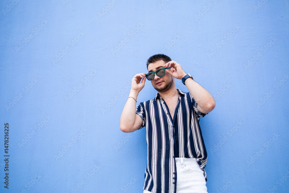 A stylish young man radiates confidence in trendy clothes and sunglasses against a striking blue wall, embodying a vibrant and playful youthful lifestyle in his captivating headshot