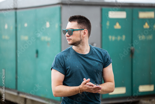 Obraz na płótnie A stylish young man wearing trendy sunglasses is confidently posing outdoors in front of green storage units on a rainy day, exuding a cool attitude and showcasing a fun personality with his apparel w salonie