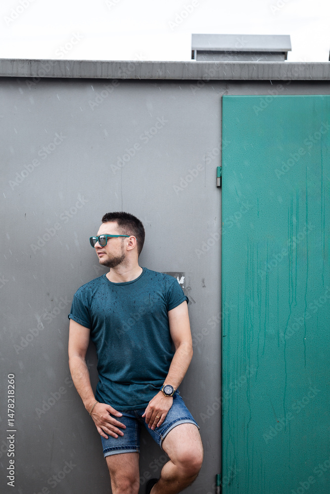 A stylish young man poses confidently against a textured wall with a vibrant green door. Dressed in trendy summer apparel and fashionable sunglasses, he radiates effortless charm and confidence