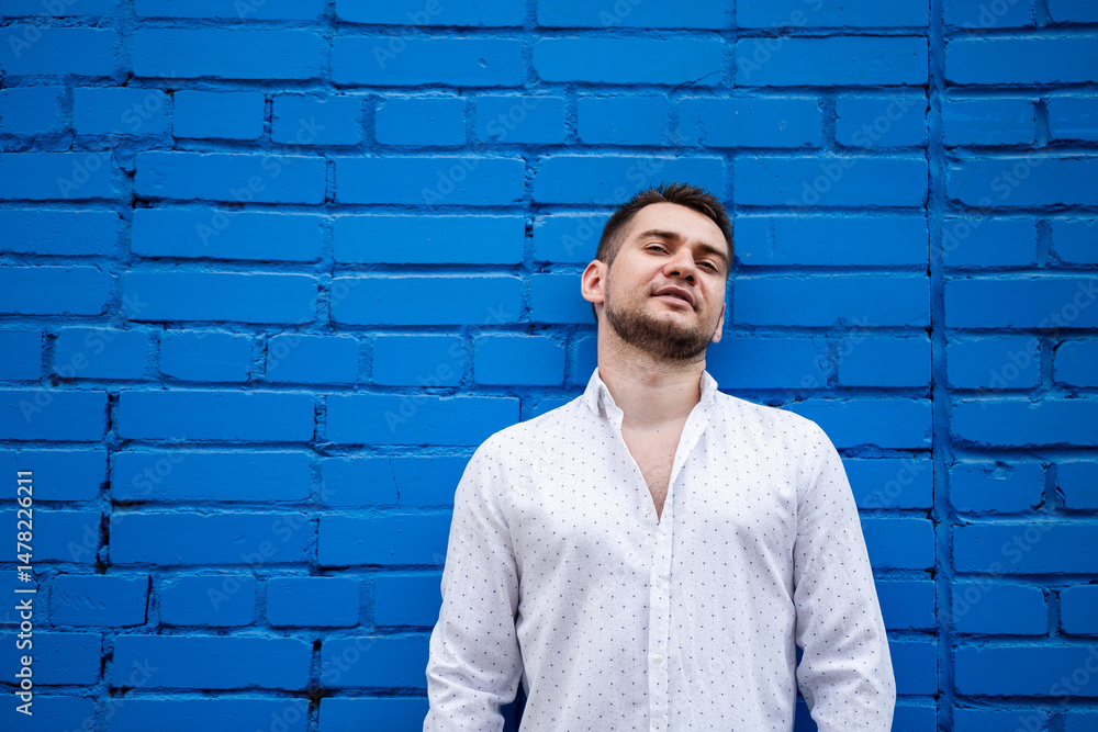 A young man elegantly posing against a bright blue brick wall while wearing a fashionable outfit. He captures a relaxed smile that beautifully reflects his individuality, charm, and youthful spirit