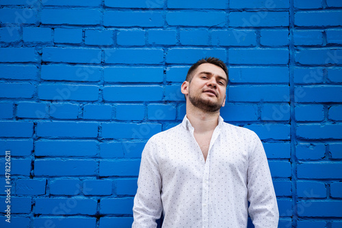 Obraz na płótnie A young man elegantly posing against a bright blue brick wall while wearing a fashionable outfit. He captures a relaxed smile that beautifully reflects his individuality, charm, and youthful spirit w salonie