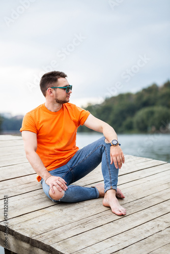 Obraz na płótnie A casually dressed young man is relaxing comfortably on a dock beside the water, wearing a bright orange tshirt and fashionable denim jeans. The scene exudes a sense of freedom and tranquility w salonie