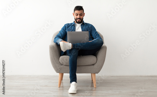 Photography Young Arab man in casual wear working online, sitting in armchair and using laptop against white studio wall, full length