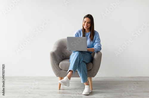 Cuadro en lienzo Happy Caucasian lady sitting in armchair with modern laptop, studying remotely or having business meeting against white studio wall, full length