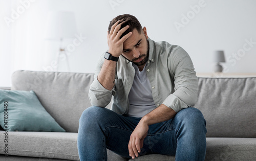 Unhappy man touching hair, sitting on sofa at home, thinking about problems. Upset guy feeling lonely, sad, have psychological and mental troubles, suffering from bad relationship or break up