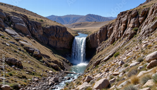 Stunning waterfall cascades between two rocky cliffs, surrounded by lush greenery and rugged terrain. serene landscape showcases beauty of nature, with mountains background and clear blue sky