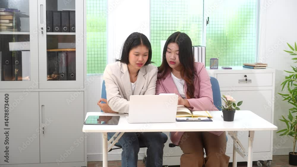 Two Asian business woman are discussing business reports and analyzing data at a desk. One is using a tablet while the other gestures. They appear focused,exchanging ideas in a modern office setting Two Asian business woman are discussing business reports and analyzing data at a desk. One is using a tablet while the other gestures. They appear focused,exchanging ideas in a modern office setting