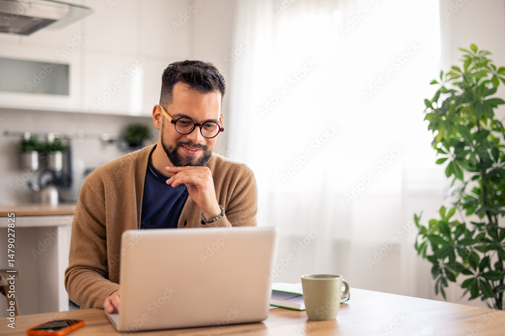 Man with Glasses Working on Laptop at Sunlit Kitchen Table with Green Plant Background Man with Glasses Working on Laptop at Sunlit Kitchen Table with Green Plant Background