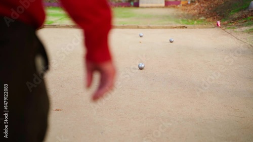 Petanque player throwing heavy metal boule with precision during competitive outdoor leisure activity, showcasing concentrated skill against sandy court background