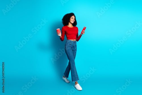 Cheerful young woman in red top holding coffee and phone against blue background embracing vibrant casual lifestyle