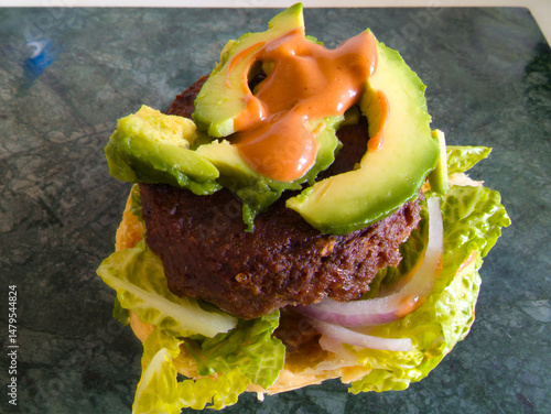 Burger with beef patty on lettuce topped with avocado slice and burger sauce served on green marble chopping board 
