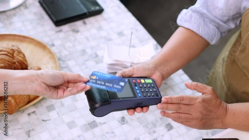 Senior entrepreneur cafe owner barista holds POS terminal while customer swipes creditcard, Asian retired female in apron smiling at counter, demonstrating cashless payment inside modern coffee shop