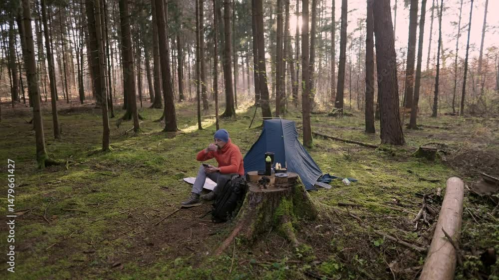 Male hiker drinking tea and surfing Internet on smartphone in tent camp during hiking in fall in woods. Backpacker makes route in map in phone and drinks hot drink from cup in background of tent. 