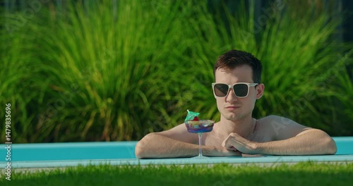 A man in sunglasses relaxes by the pool with a drink, enjoying a summer day surrounded by greenery.