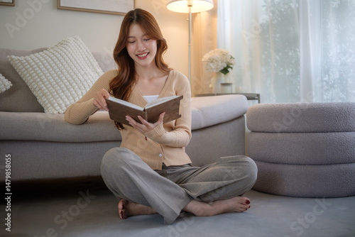 Young Asian woman sitting comfortably on the floor at home, smiling while reading a book in a cozy, softly lit living room.