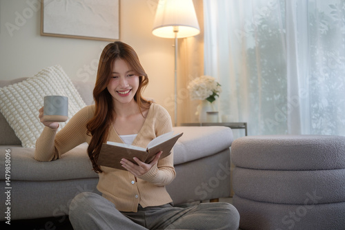 Happy young woman sitting comfortably at home, enjoying a warm drink and reading a book in a peaceful living room.