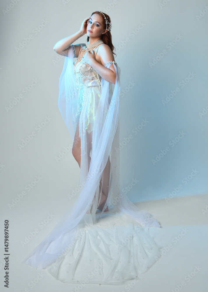Fototapeta premium Fantasy studio portrait of a red-haired mermaid faery wearing a white pearl gown, fairytale costume. posing in a classical standing position with flowing fabric and ethereal studio lighting