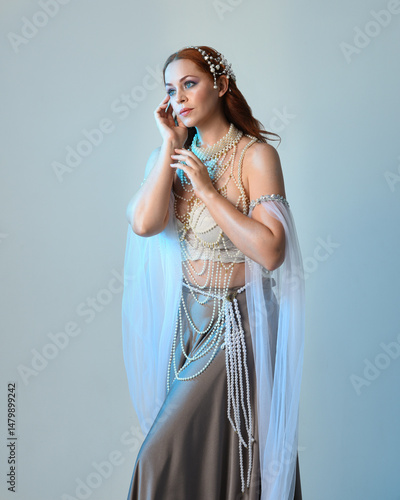 Fantasy studio portrait of a red-haired mermaid faery wearing a white pearl gown, fairytale costume. posing in a classical standing position with flowing fabric and ethereal studio lighting