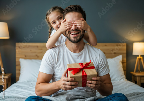 Daughter surprising her smiling dad with wrapped gift on father’s day in cozy bedroom interior