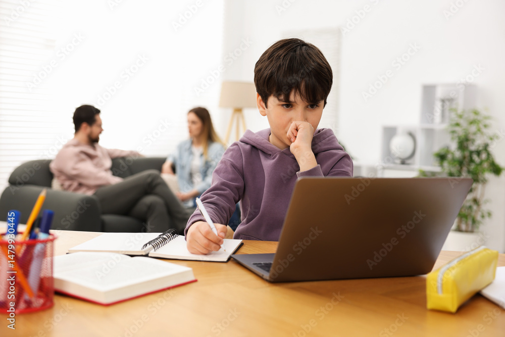 Boy doing homework at wooden table while his parents talking on sofa indoors, selective focus Boy doing homework at wooden table while his parents talking on sofa indoors, selective focus