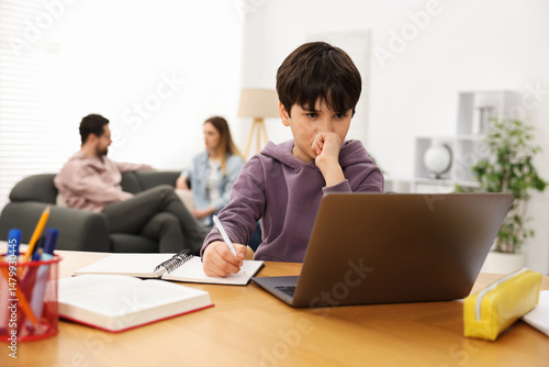 Boy doing homework at wooden table while his parents talking on sofa indoors, selective focus