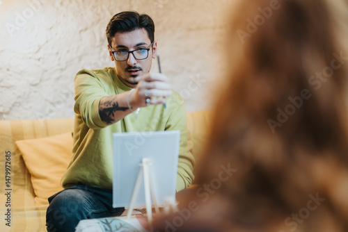Wall Mural A young artist in glasses and a tattoo, engrossed in a thoughtful artistic interaction during a painting session, instructing and guiding a subject with precision