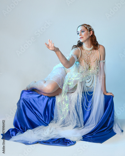 Fantasy studio portrait of a red-haired mermaid faery model wearing a white pearl gown, fairytale costume. posing in a classical sitting position with flowing fabric and ethereal studio lighting