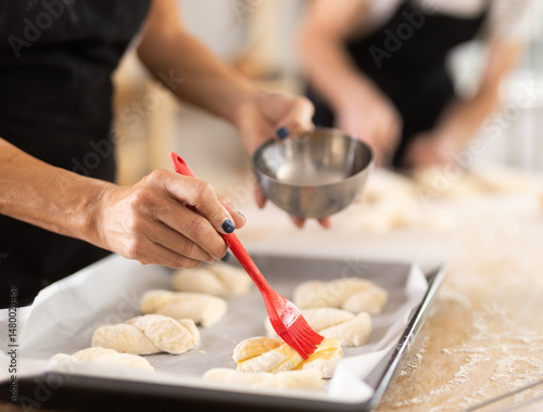 Female hands brushing uncooked dough pieces on baking sheet with oil before baking in oven closeup