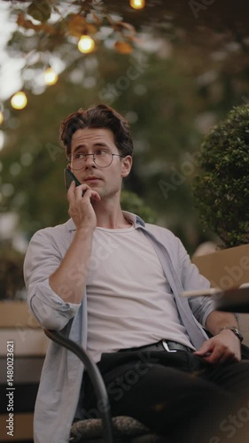 A young man sits at an outdoor cafe table, talking on his phone. He looks concerned as he listens to the person on the other end.