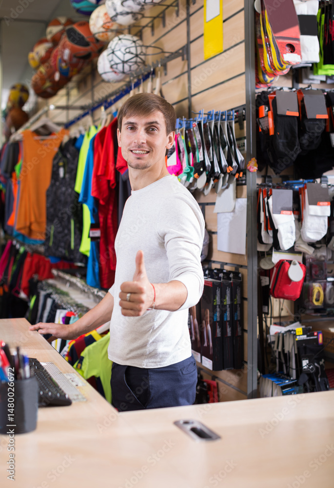Male sportswear store salesperson at the store counter. Man happy with sales - holds thumb up