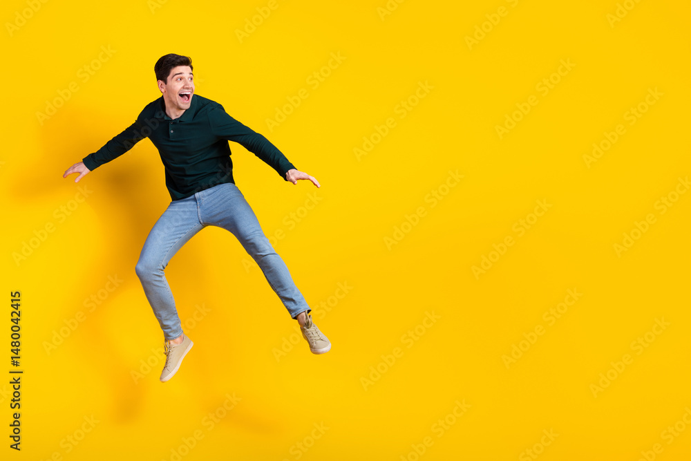 Fototapeta premium Young man jumping with joy on a yellow background wearing a green shirt and jeans showcasing happiness and vibrant energy