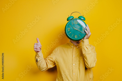 Man holding a clock in front of his head over yellow wall showing thumbs up