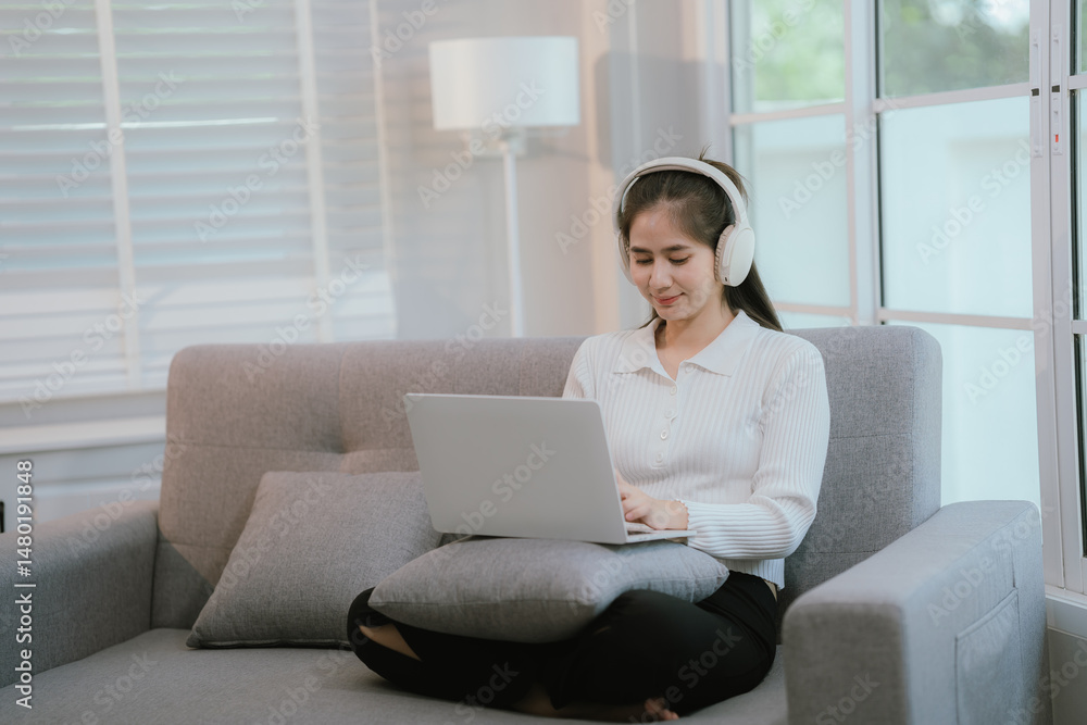 Naklejka premium Asian woman living lifestyle wearing wireless headphones staring at laptop screen and communicating with clients freely on sofa at home.
