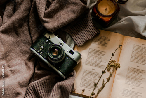 Vintage film camera with an old book wrapped in a warm knitted sweater and an extinguished candle next to it. View from above on a flat surface.