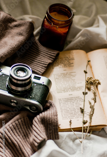 Vintage film camera with an old book wrapped in a warm knitted sweater and an extinguished candle next to it. View from above on a flat surface.