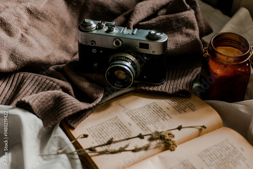 Vintage film camera with an old book wrapped in a warm knitted sweater and an extinguished candle next to it. View from above on a flat surface.