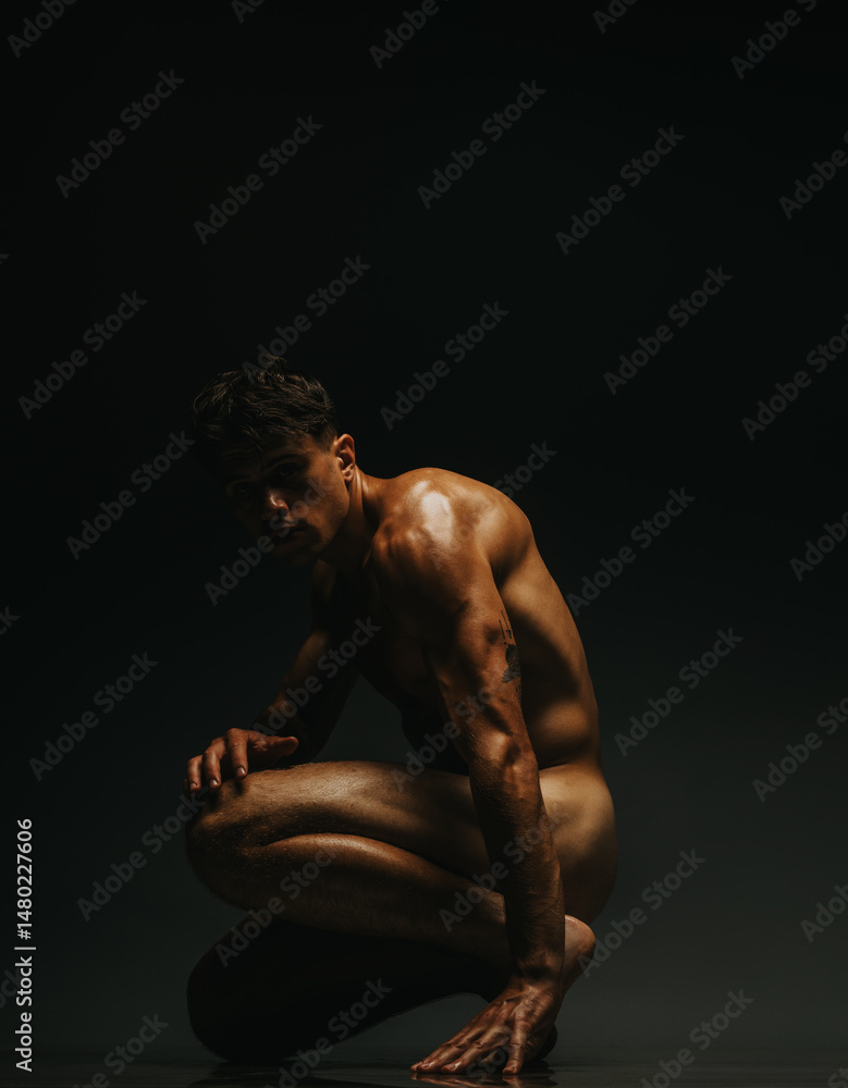Fototapeta premium A muscular man kneels in a dynamic pose, captured under low-light dramatic shadows.