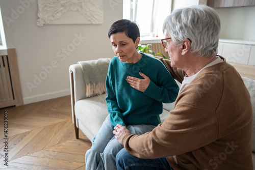 Heavily breathing woman grip chest on sofa during sudden panic attack while husband unsure how to help. Chest pain, heart attack, fear confusion and helpless in chronic illness, old couple problem