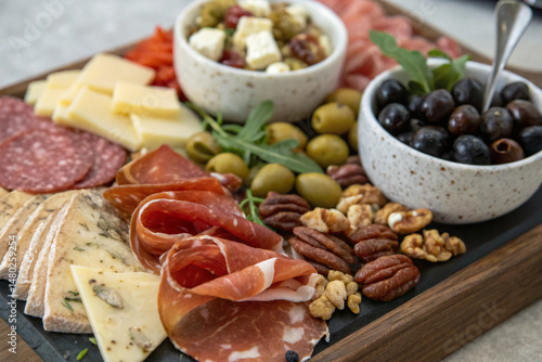 This photograph captures a beautifully arranged charcuterie and cheese board featuring salami, white and yellow cheeses, olives, almonds, and pecans on a wooden surface.