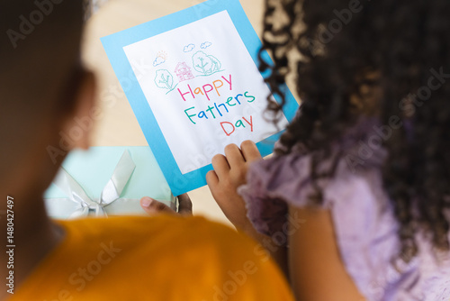 Sitting two siblings holding Happy Fathers Day card at home table, with mint-green gift box