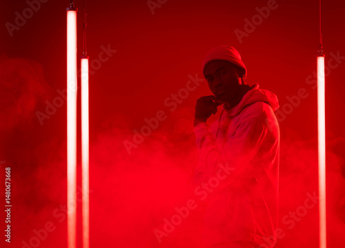 Standing African American man wearing hoodie and beanie amid red smoke and neon tubes in studio