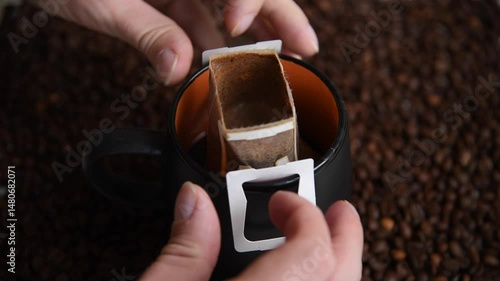 Close-up view of person taking off white paper drip coffee bag from black and orange mug with fresh brewed arabica coffee drink standing on roasted coffee beans.
