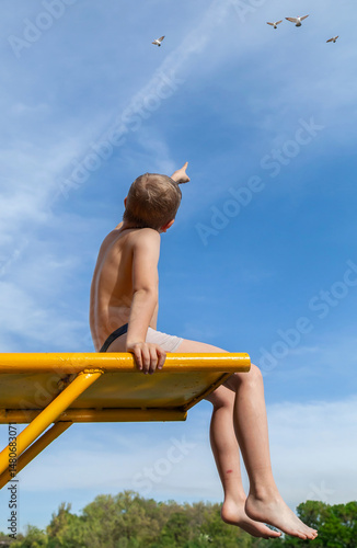 Boy sitting on bench pointing at airplane in sky. Concept of wonder and dream of flying