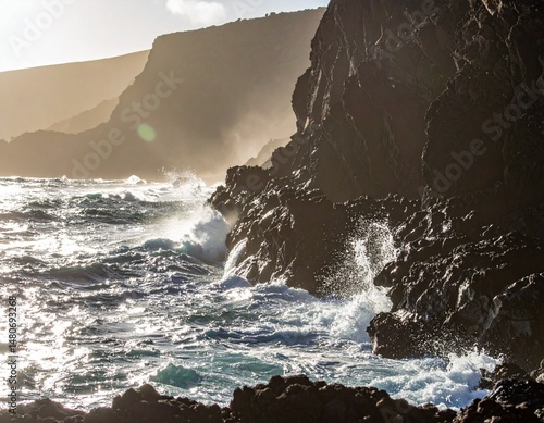 Coastal rock cliffs with waves crashing at the base, sunlit water spray, and dramatic photo