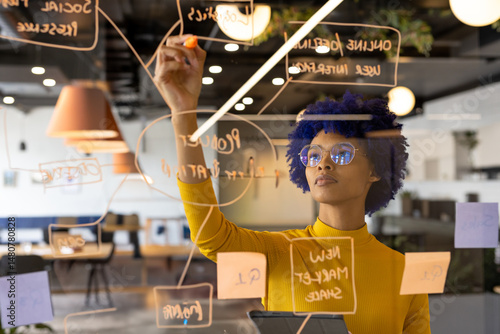 Young African American woman writing on glass board in open-plan office, with sticky notes, tablet