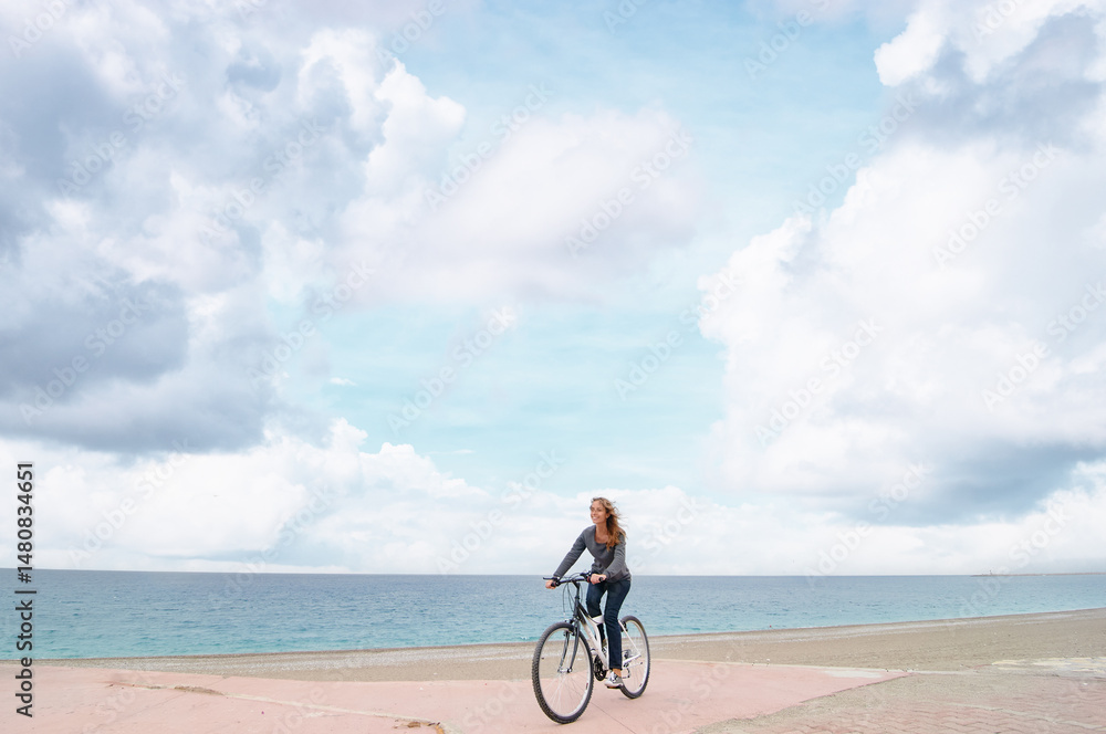 Fototapeta premium Woman Riding a Bicycle on a Beachside Path with Blue Sky and Clouds..
