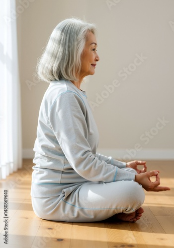 elderly Asian woman meditating in yoga pose on floor with peaceful smile in bright room