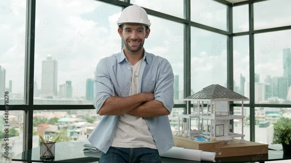 Civil engineer put on safety helmet while standing with arm folded. Professional architect engineer smiling, looking at camera while standing near house model and window with sky scraper. Tracery