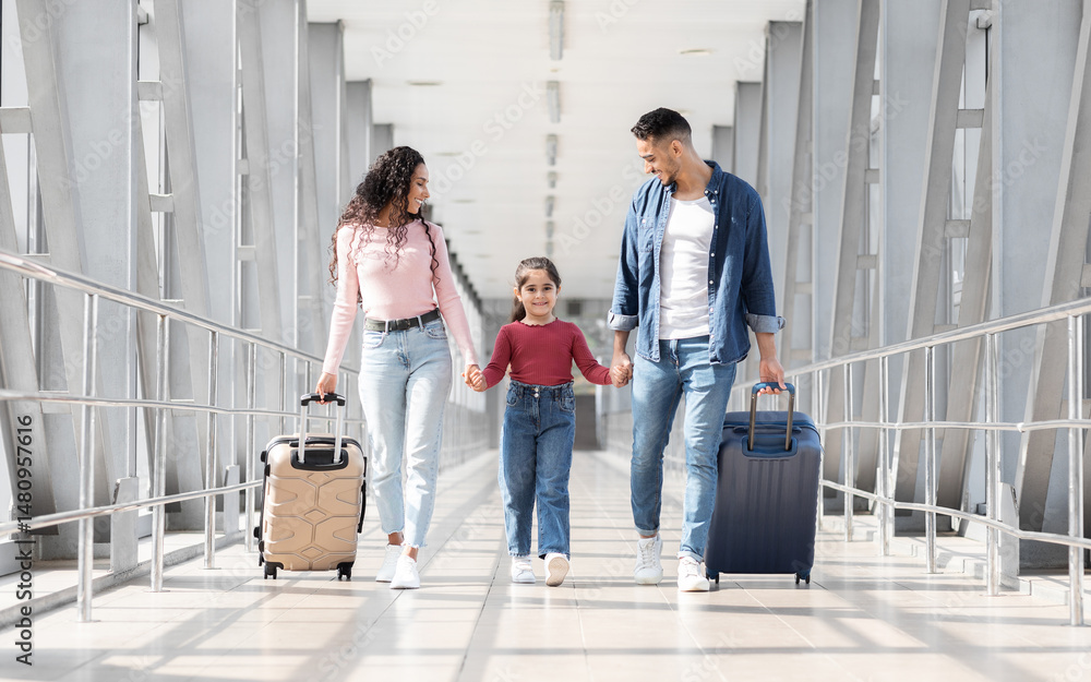 Fototapeta premium Traveling Together. Joyful Arabic Family Walking With Suitcases At Airport Terminal, Happy Middle Eastern Parents And Female Kid Carrying Luggage While Going To Flight Departure Gate, Copy Space